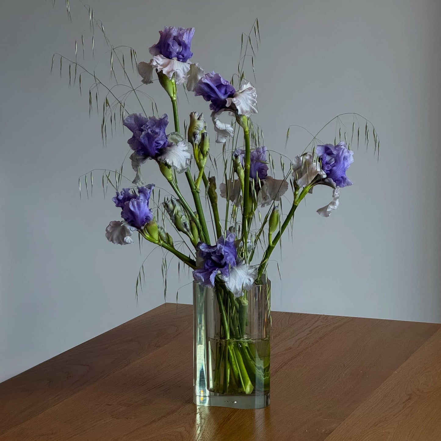 Acrylic vase with purple and white irises on a wooden table, modern floral arrangement in natural light.