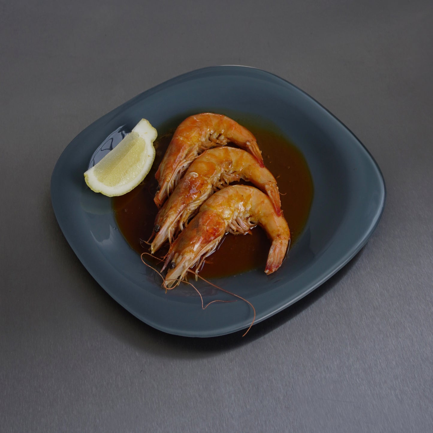 Three cooked shrimp with a lemon wedge on a grey plate against a chrome table